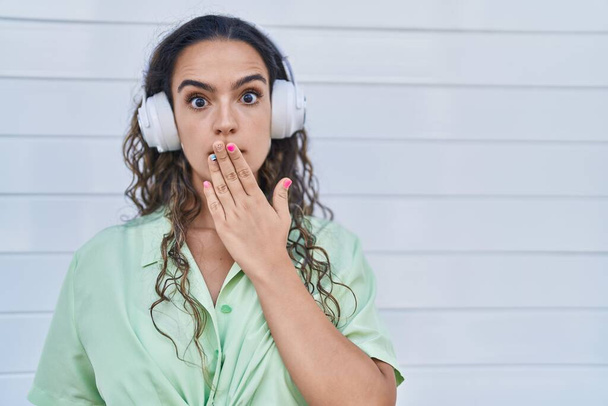 Young hispanic woman listening to music using headphones covering mouth with hand, shocked and afraid for mistake. surprised expression  - Foto, immagini