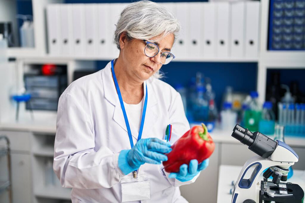 Middle age woman wearing scientist uniform analysing pepper at laboratory - Photo, image