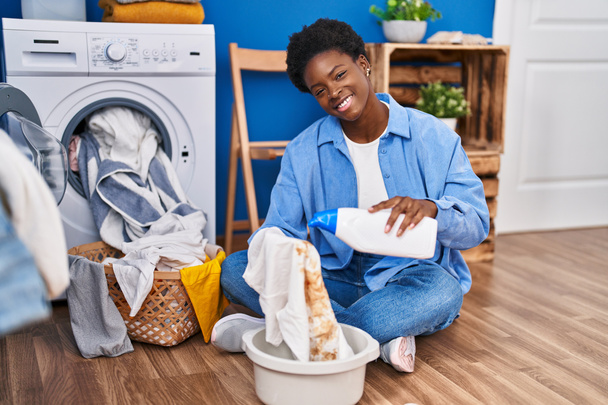 アフリカ系アメリカ人女性が洗濯室で汚れたTシャツに自信を持って洗剤を注ぐ笑顔 - 写真・画像