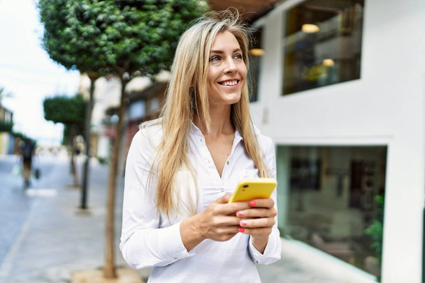 Beautiful blonde woman smiling happy outdoors on a sunny day using smartphone - Foto, afbeelding