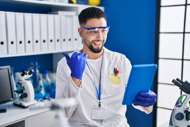 Young handsome man working at scientist laboratory doing online call pointing thumb up to the side smiling happy with open mouth  - Photo, Image