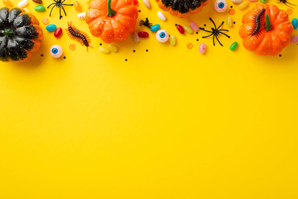 Top view photo of halloween decorations pumpkins spooky eyes centipedes candies spiders and confetti on isolated yellow background with copyspace - Foto, afbeelding