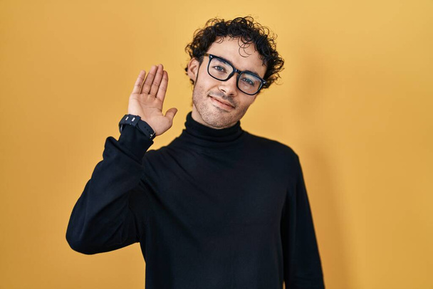 Hispanic man standing over yellow background waiving saying hello happy and smiling, friendly welcome gesture  - Photo, Image