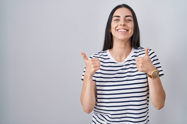 Young brunette woman wearing striped t shirt success sign doing positive gesture with hand, thumbs up smiling and happy. cheerful expression and winner gesture.  - Foto, immagini