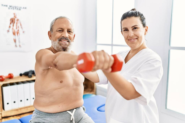 Физиотерапевт и пациент уверенно улыбается, имея сеанс реабилитации с использованием гантели в клинике - Фото, изображение