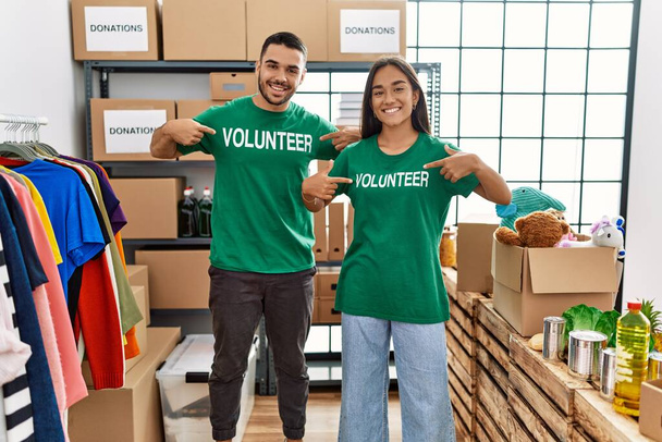 Νεαρό ζευγάρι Λατίνων χαμογελά ευτυχείς δείχνοντας εθελοντής στολή στο κέντρο φιλανθρωπίας. - Φωτογραφία, εικόνα