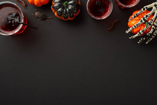 Halloween concept. Top view photo of blood punch in glasses with floating spiders and straw skeleton hand holding pumpkin centipedes and cockroach on isolated black background with copyspace - 写真・画像