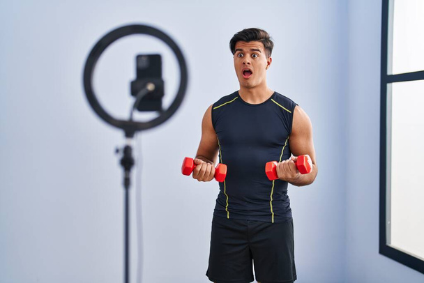 Hispanic man recording training tutorial at the gym in shock face, looking skeptical and sarcastic, surprised with open mouth  - Photo, Image