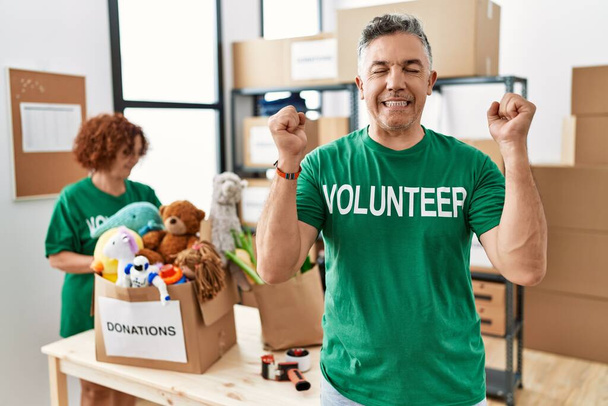 Középkorú férfi visel önkéntes póló adományok állnak izgatott siker karok fel, és csukott szemmel ünnepli győzelem mosolyogva. győztes koncepció.  - Fotó, kép