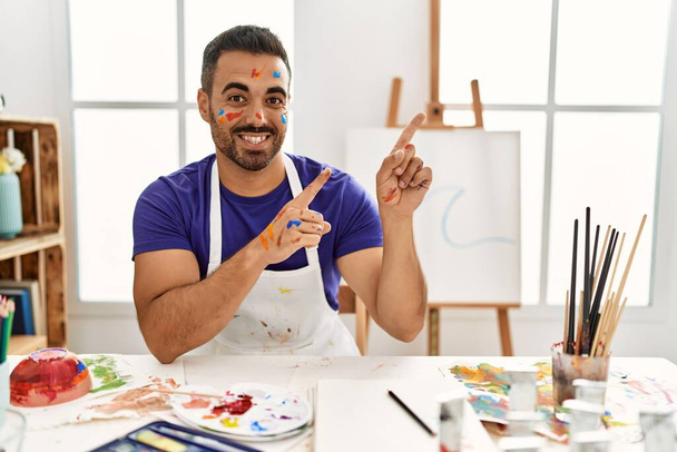 Young hispanic man with beard at art studio with painted face smiling and looking at the camera pointing with two hands and fingers to the side.  - Fotografie, Obrázek
