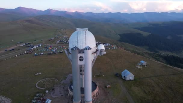 Two large telescope domes at sunset. Drone view of Assy-Turgen Observatory. Beautiful red sunset. Green hills and clouds. Tourists watch the sun. There is a large tent camp and cars nearby. Kazakhstan - Video