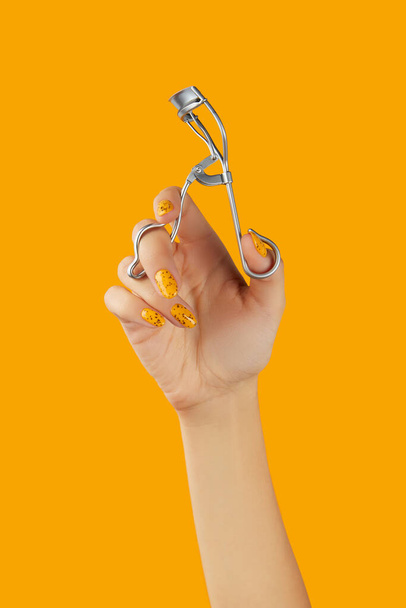 Eyelash curler in females hand on orange background. Beauty salon concept - Foto, imagen