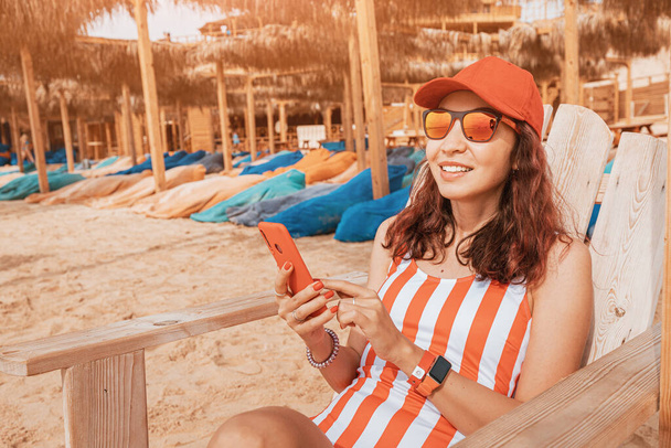 Счастливая девушка читает новости в социальных сетях в своем смартфоне, загорая на лежаке на курорте - Фото, изображение