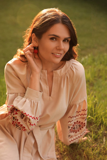 Beautiful woman in embroidered dress sitting on green grass outdoors. Ukrainian national clothes - Foto, Imagem