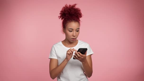 African American shocked woman with red hair using smartphone typing browsing on isolated pink background. African female wearing white T-shirt holding mobile phone and is surprised by big online sale - Video
