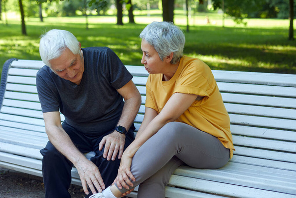 During morning stroll, sportive exercises in park, elderly 60s woman got injured ankle, grip foot sit in bench with caring disappointed husband. Traumas, injures of older people concept - Foto, Imagen