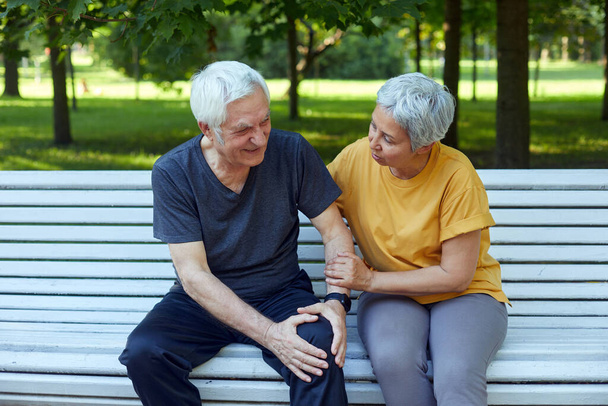 During morning sportive stroll or making exercises in a park, elderly 60s man got injured his knee, gripping leg sit in bench with caring disappointed wife. Traumas, injures of older people concept - Foto, immagini