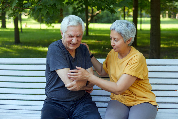 During morning sportive stroll or making exercises in a park, elderly 60s man got injured his shoulder gripping arm sit in bench with caring disappointed wife. Traumas, injures of older people concept - Φωτογραφία, εικόνα
