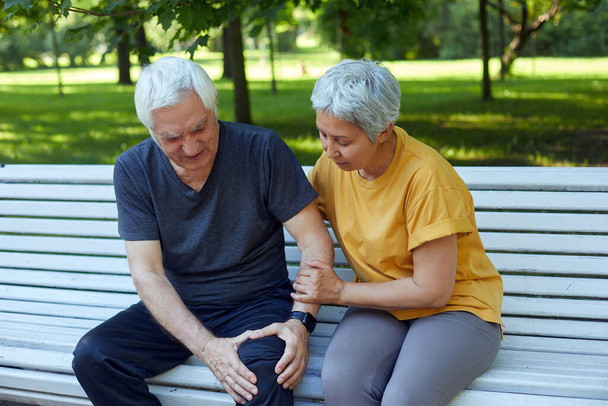 During morning sportive stroll or making exercises in a park, elderly 60s man got injured his knee, gripping leg sit in bench with caring disappointed wife. Traumas, injures of older people concept - Foto, immagini