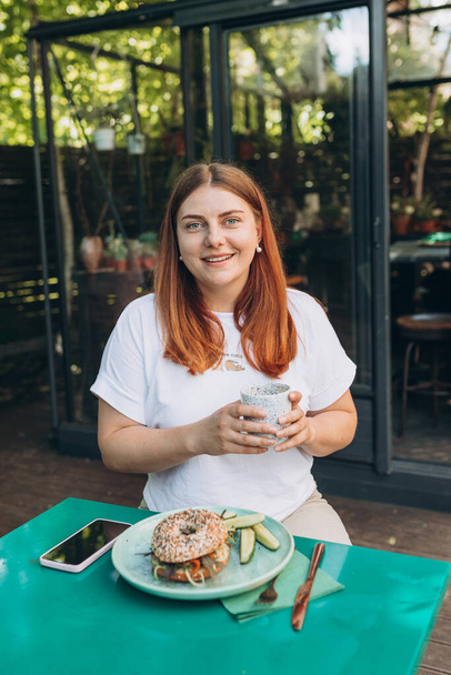 Happy Young redhead woman enjoying a coffee, sitting on the cafe terrace on the city street. Person sitting at restaurant table and drinking cappuccino in cup, Concept of easy breakfast - Fotografie, Obrázek