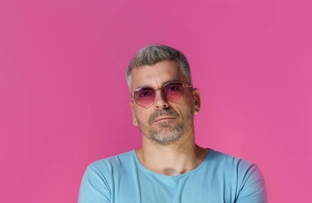 Handsome man 30s 40s wearing pink sunglasses and blue t-shirt with pensive, concentrated look isolated on pink background. Grey haired mature man studio shot.  - Foto, Imagen