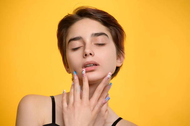 Romantic studio portrait of young pretty girl pouching her lips, short haircut and extravagant nail art, eyes closed, mouth slightly open, yellow colored background - Valokuva, kuva