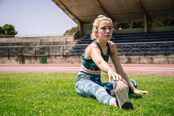 Все тело спортсменки касаясь ноги и глядя в сторону, сидя на газоне во время тренировки на легкоатлетическом стадионе летом - Фото, изображение