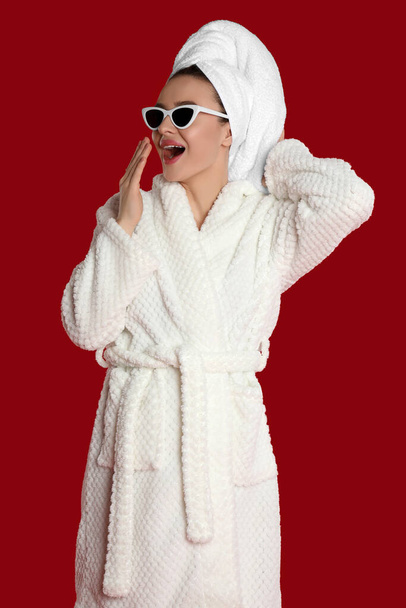 Beautiful young woman in bathrobe and sunglasses yawning on red background - Φωτογραφία, εικόνα