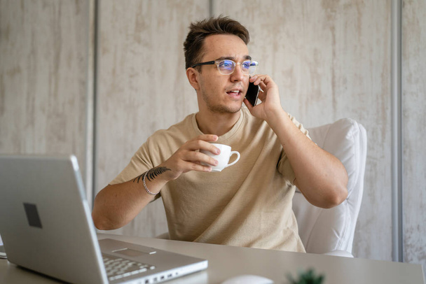 One man caucasian male sitting in chair in the office at work at desk using mobile phone to make a call while working on project wear eyeglasses talking consulting copy space real people - Foto, imagen