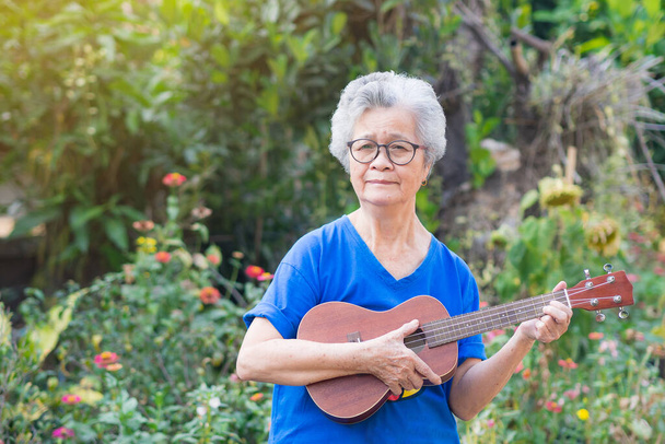 Elderly Asian woman with short gray hair, wearing glasses, playing the ukulele, and looking at the camera while standing in a garden. Space for text. Concept of aged people and relaxation. - Foto, imagen