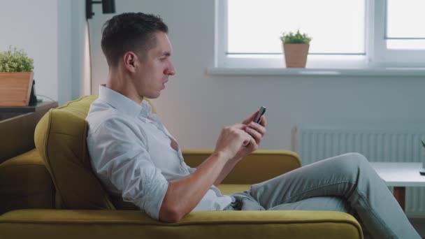 Surprised young man winner hold smartphone read good news amazed by mobile online bet bid game win at home. Happy male look at cell phone screen overjoyed by victory success while sitting on sofa. - Footage, Video