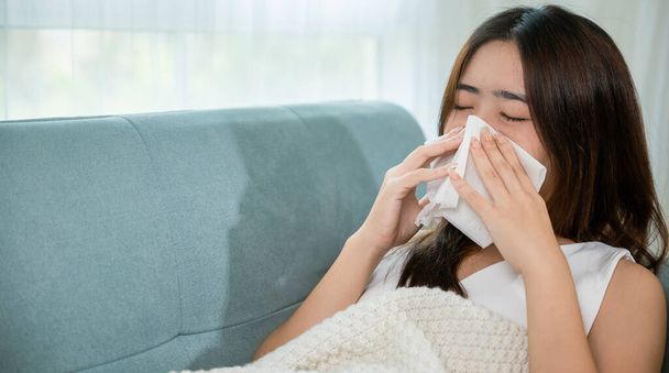 Ill Asian young woman cold covered with blanket sitting on sofa and sneeze with tissue paper at home, Female sick allergic blowing nose sneezing in tissue at home, Flu health care - Фото, зображення