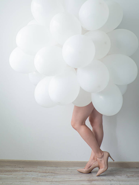Woman in beige high heels holding white color balloons on a white wall background. Legs and a lot of white air balloons. Happy birthday anniversary. White decoration. Copy space - Φωτογραφία, εικόνα