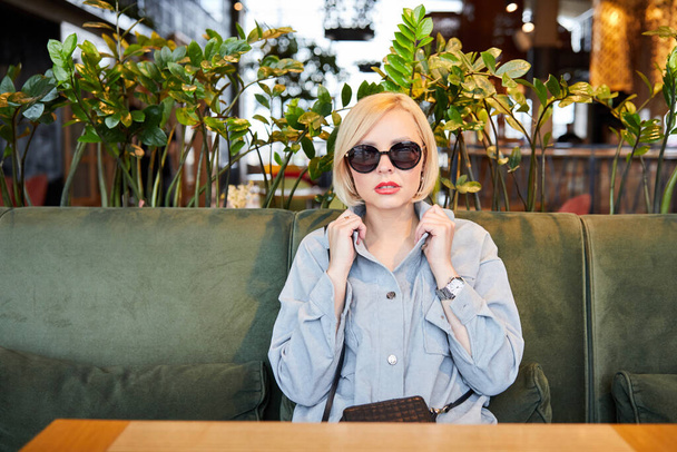 Κομψή νεαρή ξανθιά με γυαλιά ηλίου κάθεται σε ένα τραπέζι σε ένα όμορφο εστιατόριο περιμένει για μια παραγγελία. - Φωτογραφία, εικόνα