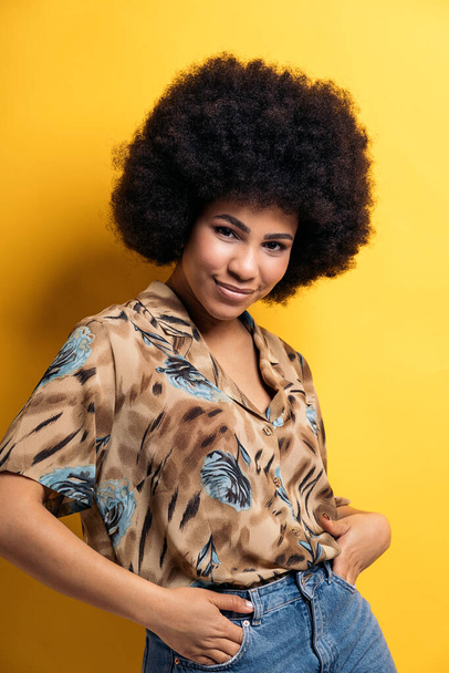 Smiley afro woman wearing colorful shirt smiling and looking at camera in studio shot against yellow background. - Фото, изображение