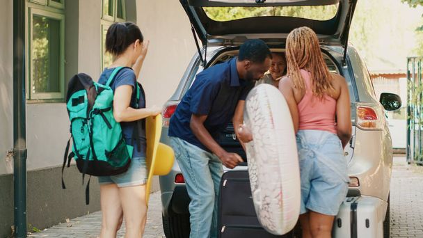 Multiethnic people loading car trunk with voyage luggage while little girl hurrying them. Diverse couple with mother and daughter putting baggage inside vehicle while going on holiday summer citybreak - Photo, image