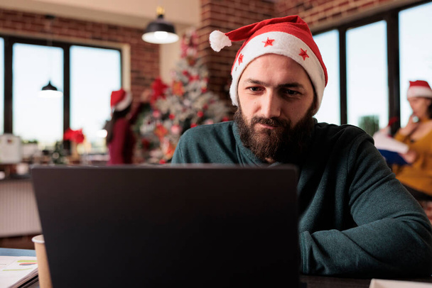 Startup employee using laptop in business office to work on company project during winter season. Man with santa hat working on report in workplace with christmas tree and holiday decorations. - Φωτογραφία, εικόνα