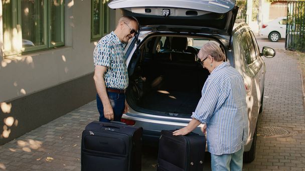 Cheerful elders going on retirement voyage while having heavy baggage and travel trolley. Senior couple putting luggage in car trunk while getting ready for holiday trip. - 写真・画像