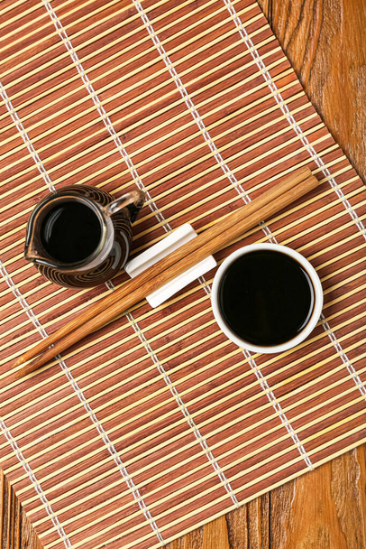 Jug and bowl of soy sauce, chopsticks and bamboo mat on wooden background - Valokuva, kuva