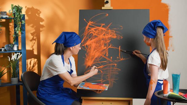 Η μαμά και το παιδί μαθαίνουν να ζωγραφίζουν με πορτοκαλί ακουαρέλα και πινέλο, δημιουργώντας σχέδια μαζί σε καμβά. Ευτυχισμένοι άνθρωποι ζωγραφική αριστούργημα με ακουαρέλα και βούρτσα. - Φωτογραφία, εικόνα