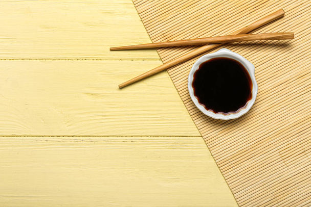 Schaal van sojasaus, stokjes en bamboe mat op gele houten ondergrond - Foto, afbeelding