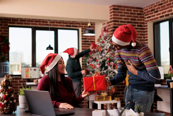 African american employee giving present to colleague, celebrating holiday season and christmas eve in festive office with seasonal decorations and ornaments. People feeling happy about xmas time. - Foto, Imagen