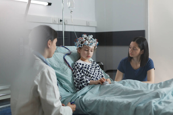 Ειδικός νευρολόγος παιδιατρικού νοσοκομείου που εξετάζει νοσοκομειακή εγκεφαλική κατάσταση κοριτσιού. Ειδικός ιατρός που μετρά την εξέλιξη νευρολογικής νόσου του άρρωστου παιδιού που κάθεται σε παιδιατρική μονάδα υγειονομικής περίθαλψης. - Φωτογραφία, εικόνα