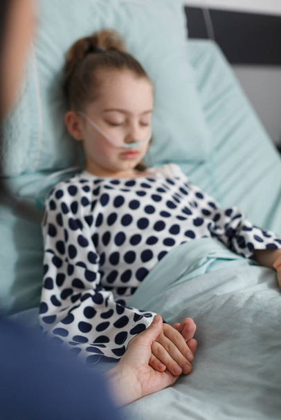 Госпитализированная больная девочка спит на больничной койке, пока мать держит ее за руку. Нездоровый ребенок отдыхает в больничной педиатрической палате, пока родители утешают ее.. - Фото, изображение