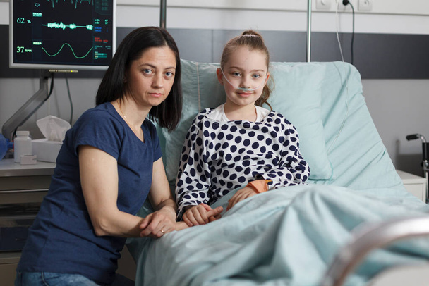 Woman sitting beside ill little girl laying in children healthcare facility room while looking at camera. Portrait shot of caring mother comforting hospitalized sick daughter resting in patient bed. - Foto, afbeelding