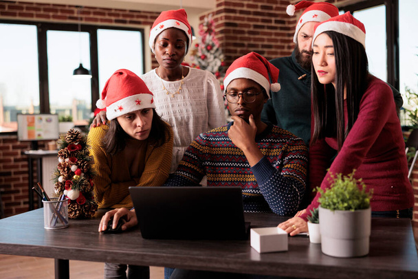 Разнообразная команда сотрудников, использующих ноутбуки в праздничном офисе, работает над запуском бизнеса во время рождественских праздников. Работа в команде на рабочем месте, украшенная рождественскими орнаментами. - Фото, изображение