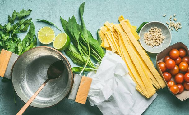 Wild garlic noodles ingredients. Fettuccine, ramson leaves,tomatoes, pine nuts, lemon and cooking pan on blue kitchen table background, top view. Seasonal food concept - Foto, imagen