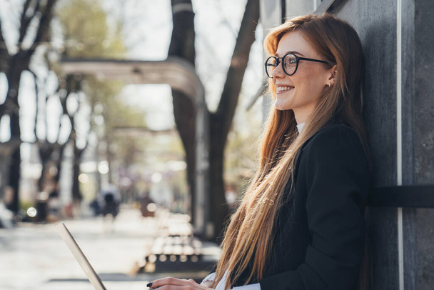 Red-haired casual woman with freckles is resting on a bench in the city park, on a summer day, using laptop. People urban lifestyle concept - Photo, image