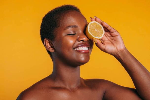 Красивая африканская американка делает маску для глаз из лимонов, позирующих на оранжевом фоне. Концепция велнес и скинкаре. Молодая черная леди держит цитрусовые - Фото, изображение