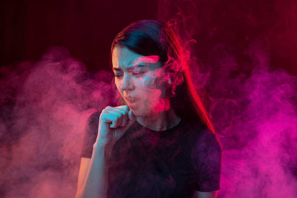 Smokers cough. Sick young man with stressed emotions isolated over dark pink background with clouds of smoke in neon. Concept of health, addiction, human emotions, challenges, ad - Photo, Image